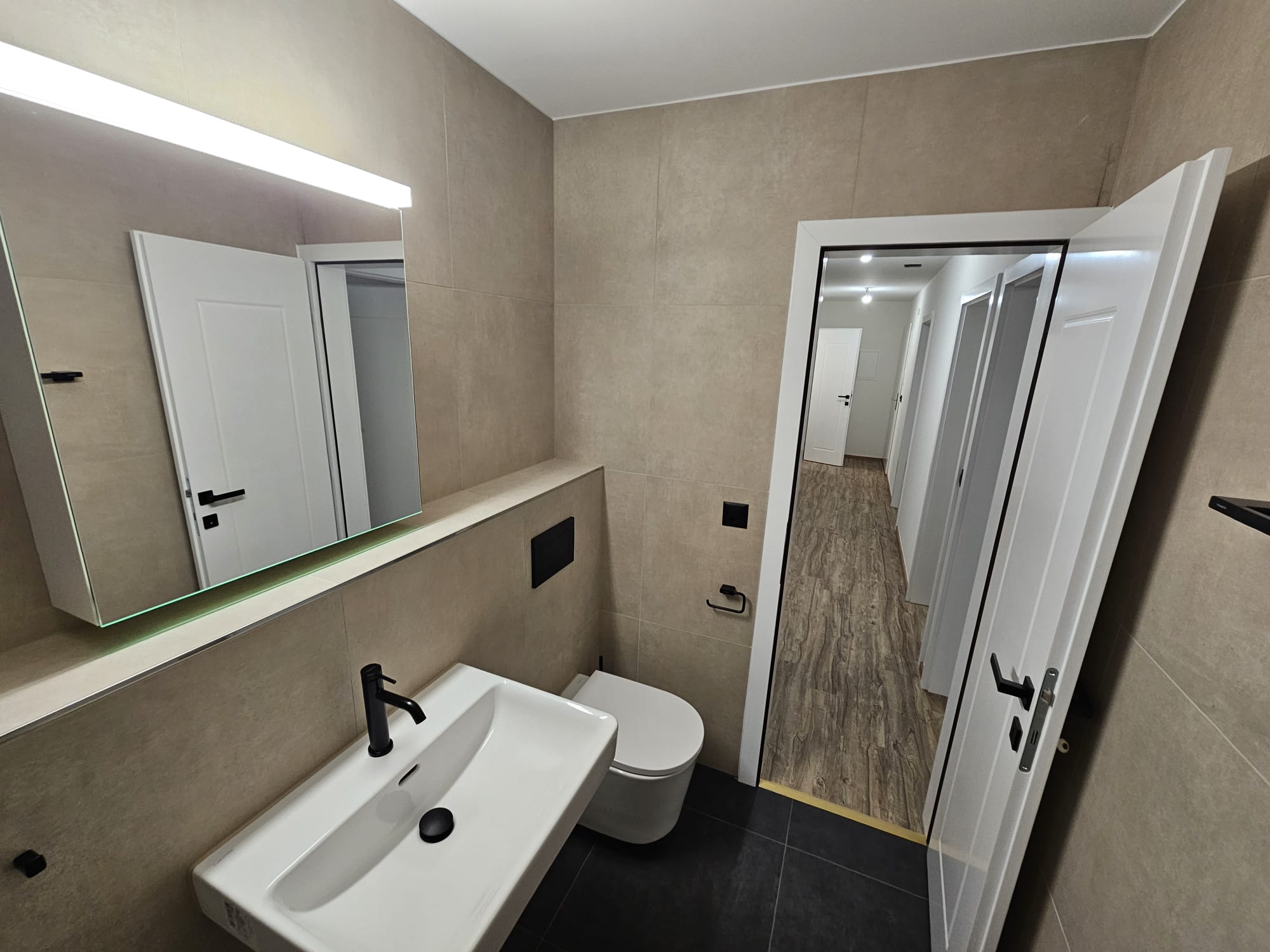 WC suspendu et lavabo : faïence beige grand format toute hauteur, sol anthracite et seuil raccordé au sol effet bois du couloir