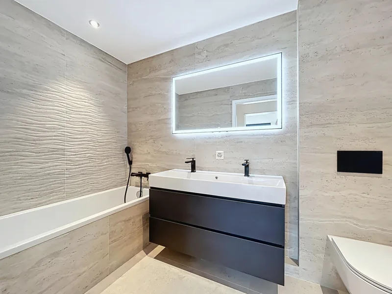 Salle de bain rénovée : baignoire sous un mur à relief ondulé, meuble vasque noir à deux robinets et miroir rétroéclairé
